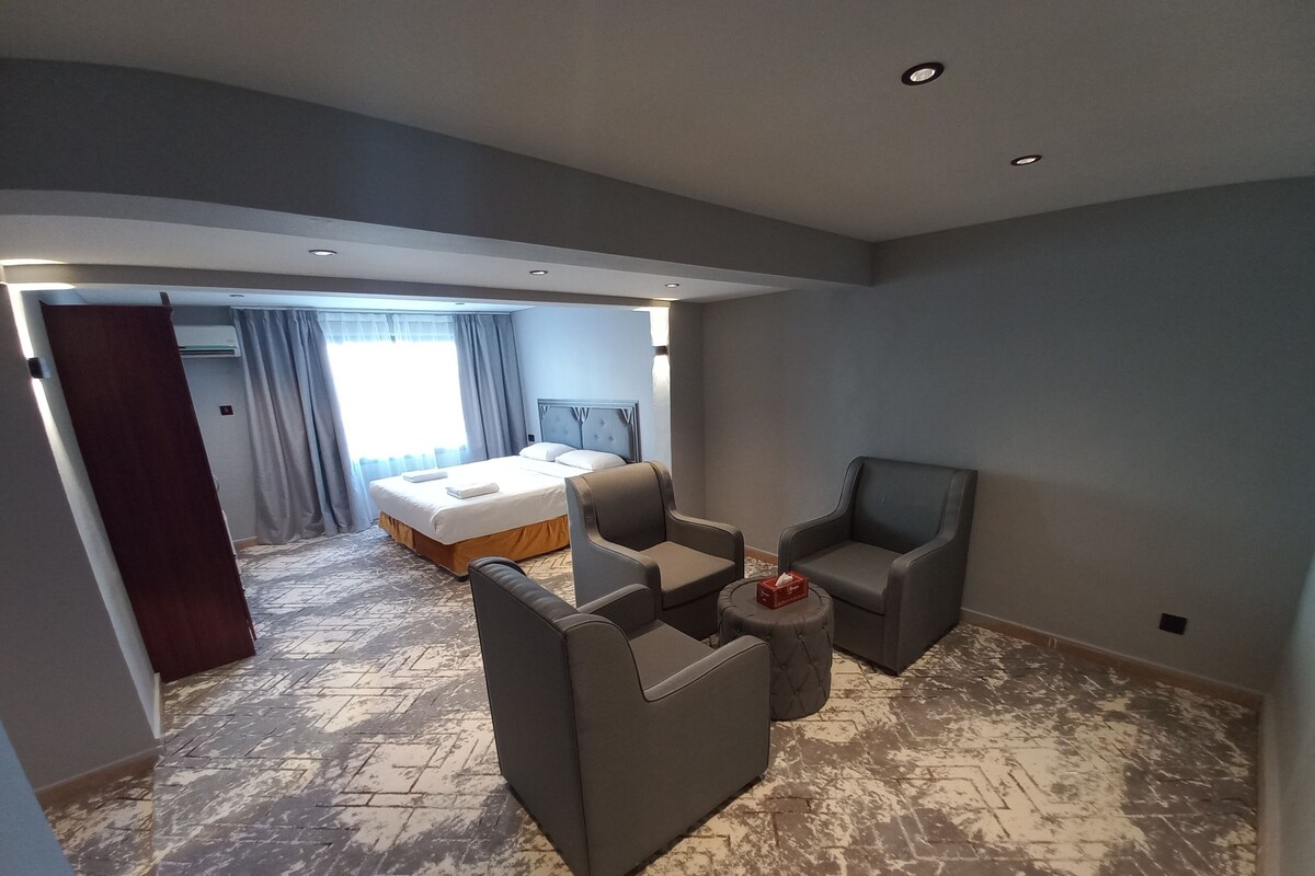 Une chambre d’hôtel moderne avec un grand lit, deux fauteuils, un petit pouf et une vue sur le couloir.