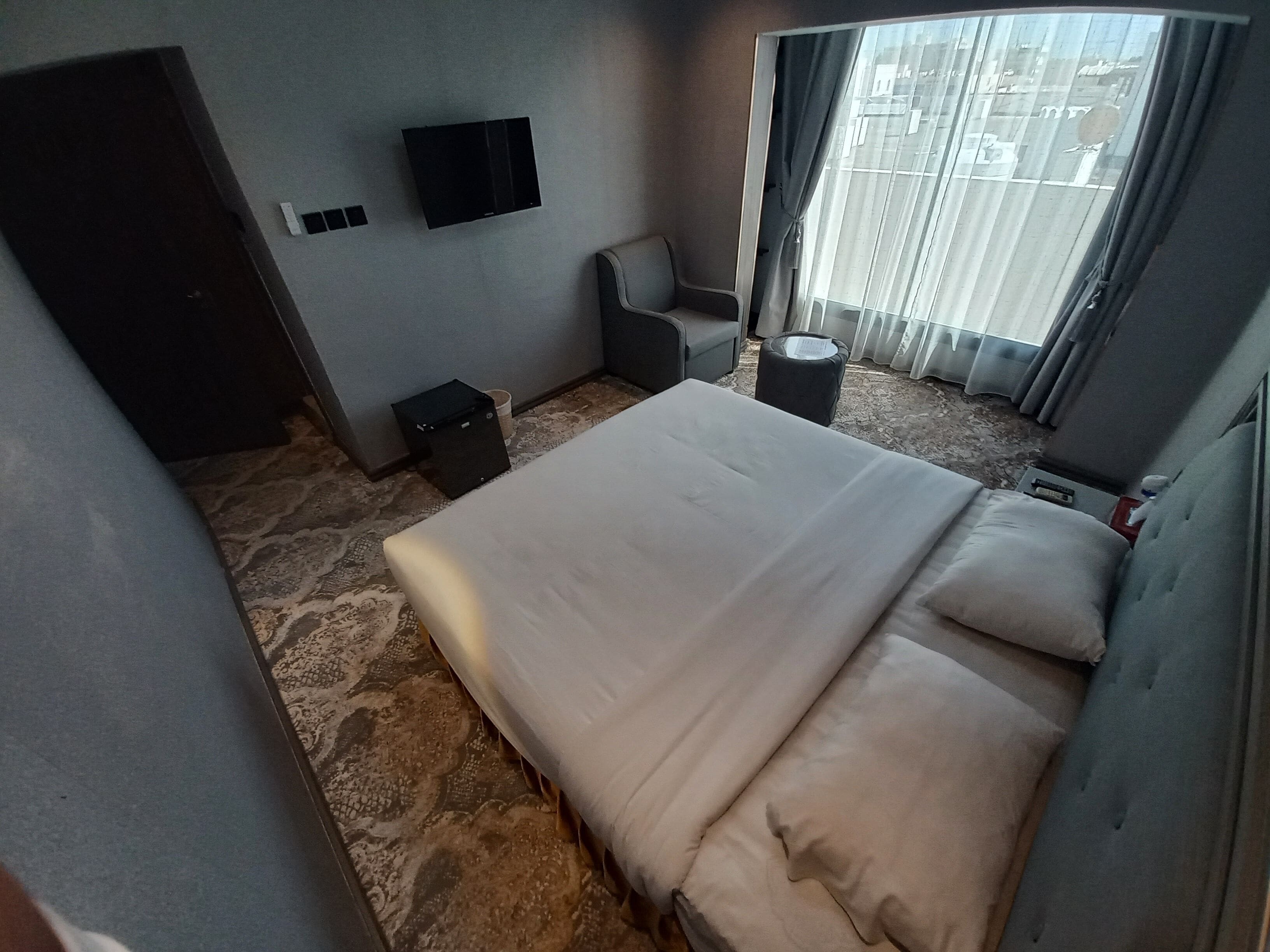 A hotel room with a bed, a chair, a small table, a trash bin, a TV mounted on the wall, and a window with curtains.