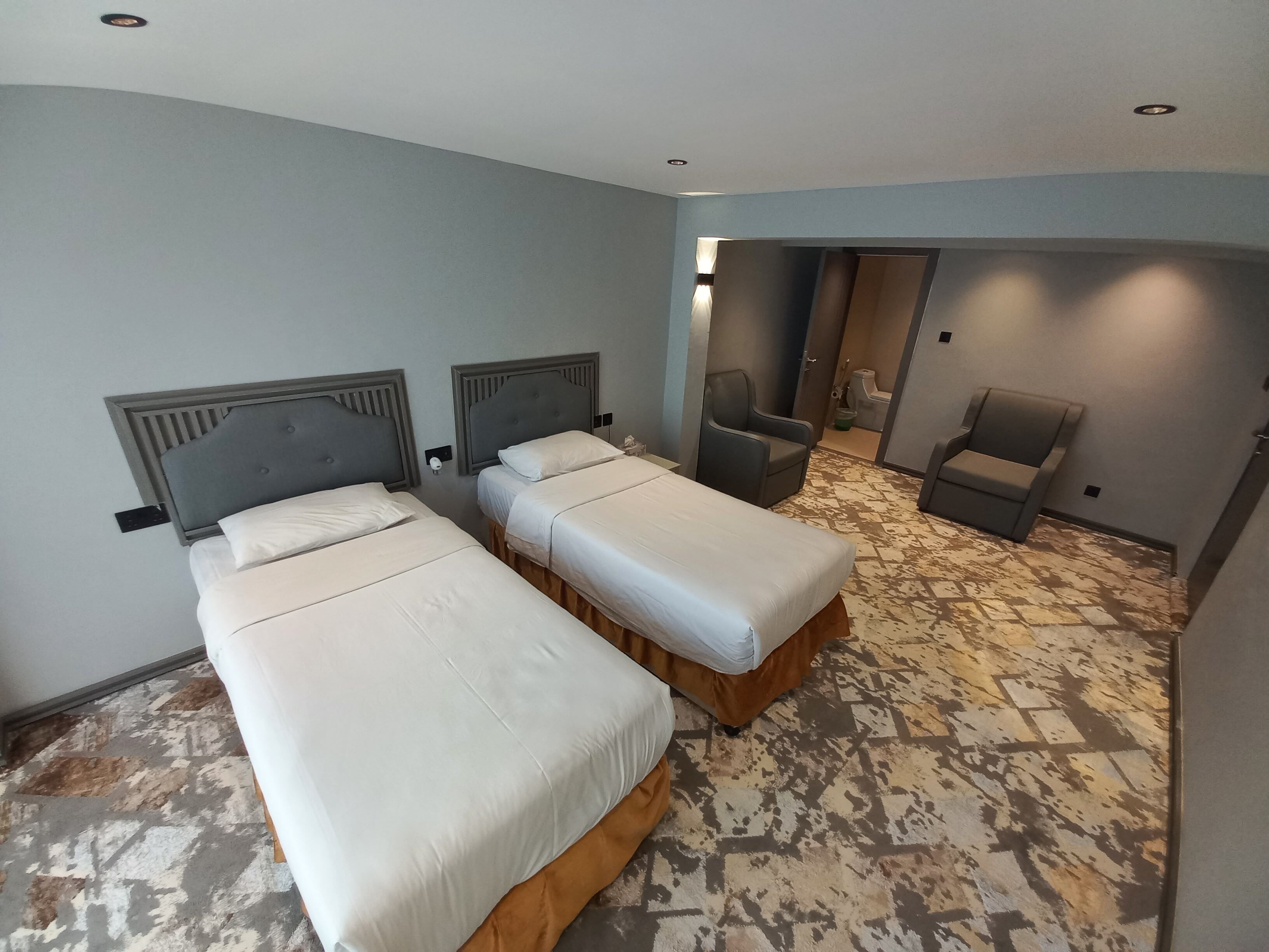 Habitación de hotel con dos camas, una silla y un baño visible a través de una puerta abierta.