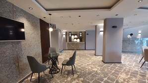 A modern hotel lobby with a marble floor, a round glass table with chairs, a flat-screen TV mounted on the wall, and a reception desk in the background.