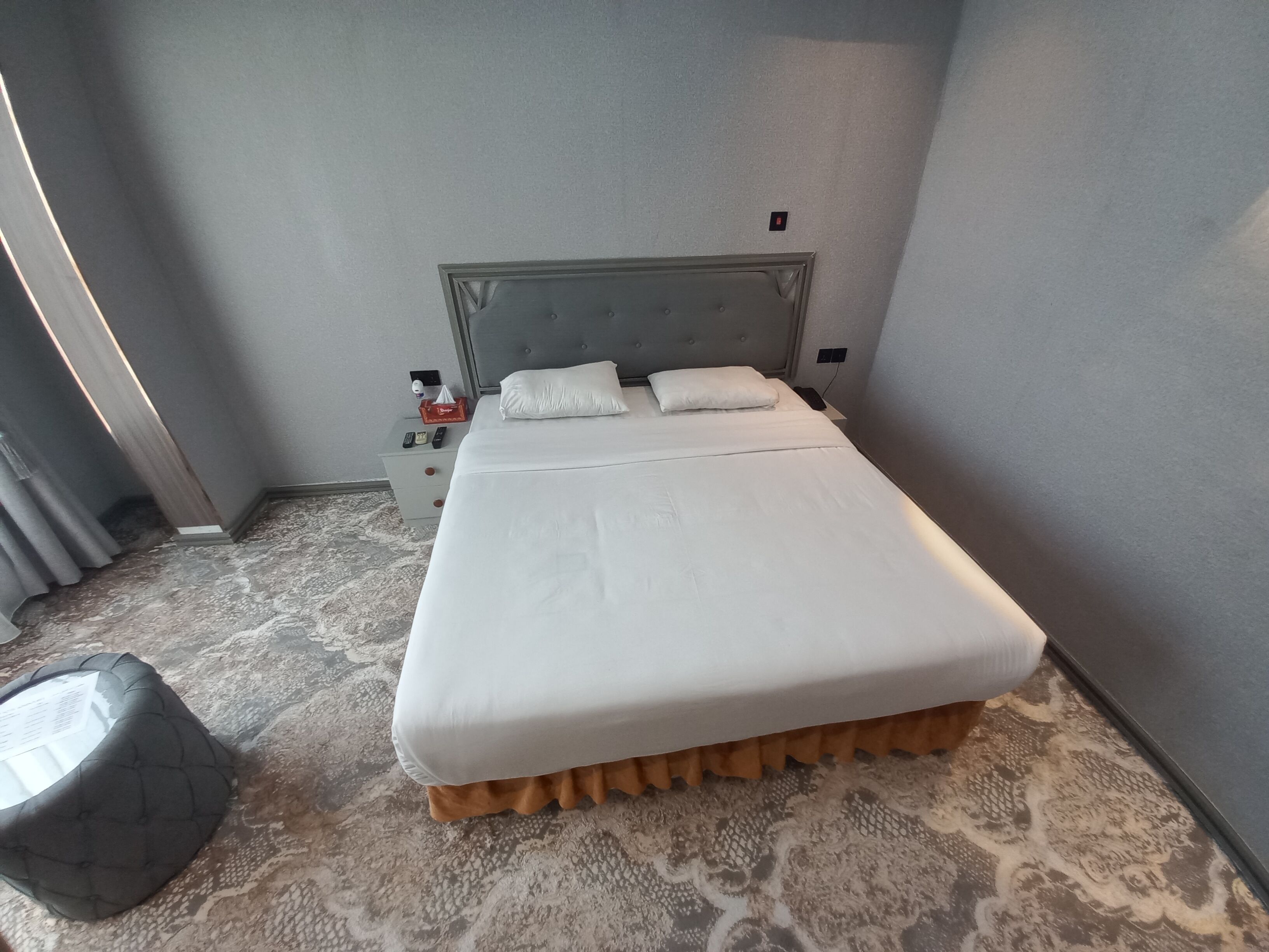 Ein ordentlich bezogenes Bett mit weißer Bettwäsche, einem grauen Kopfteil und einem kleinen Nachttisch mit Fernbedienung und Taschentuchbox.