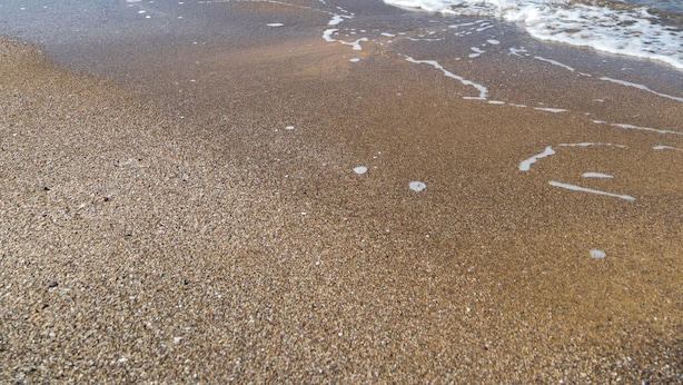 Una playa con agua azul cristalina, arena pedregosa y un acantilado rocoso al fondo.