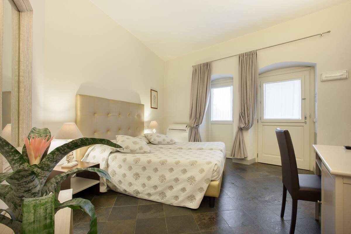 Ein Hotelzimmer mit zwei Betten, einem Schreibtisch, einem Sessel, einer Zimmerpflanze und einem Fenster mit Vorhängen.
