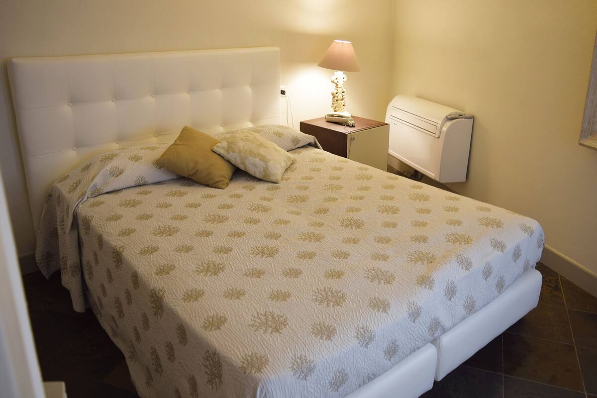 Ein ordentlich bezogenes Bett mit einer gemusterten Tagesdecke, einer Nachttischlampe und einem kleinen Nachttischchen mit Lampe.