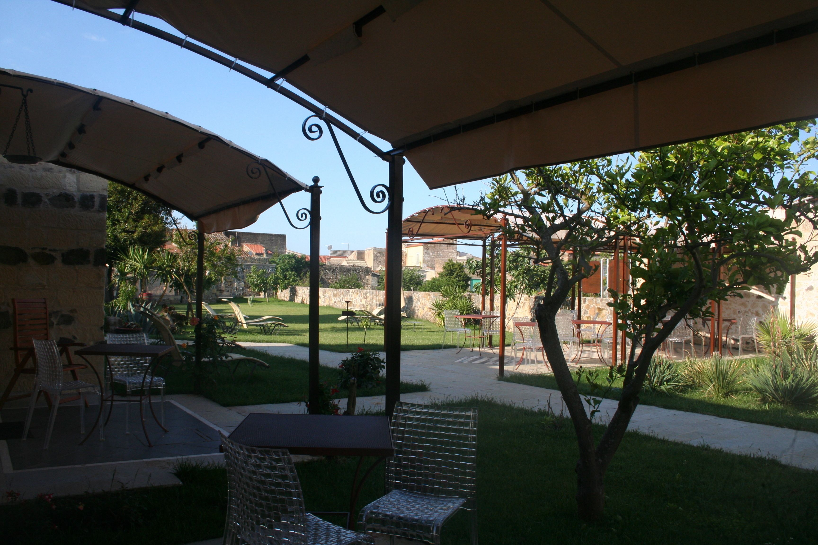 An outdoor patio area with tables and chairs under a pergola, surrounded by greenery and a view of buildings in the background.