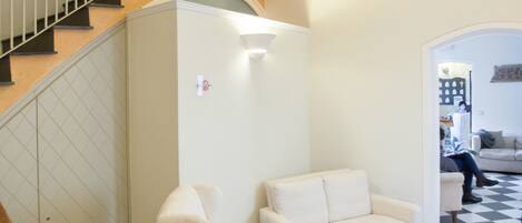 Una habitación con piso a cuadros, un sillón blanco, un sofá blanco, una escalera con barandal de madera y una puerta blanca.