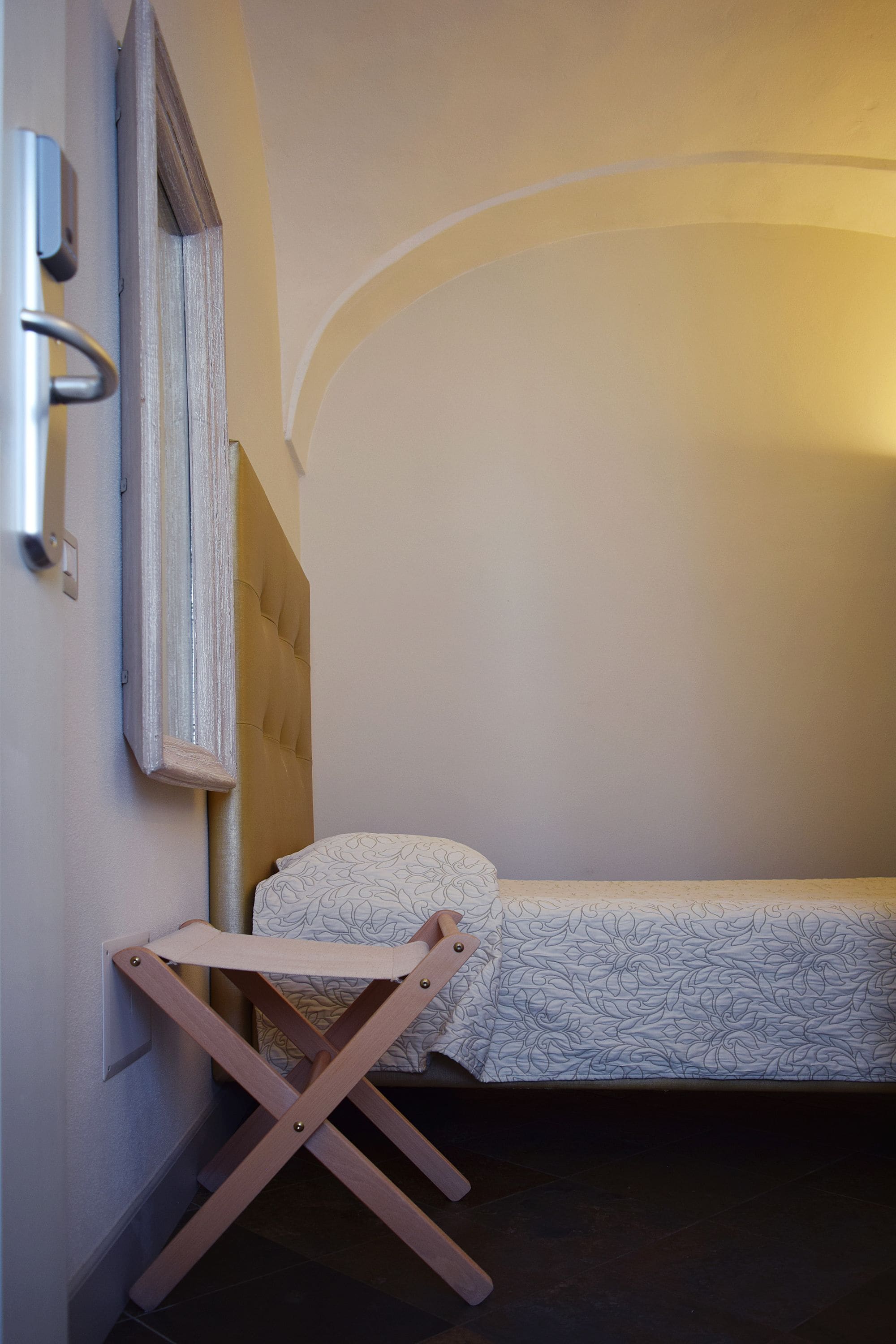 A small, neatly arranged room with a single bed, a wooden folding chair, and a wall-mounted light fixture.