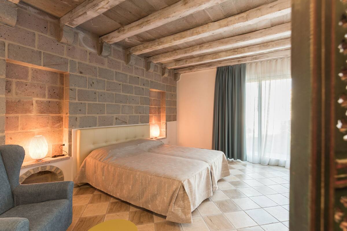 Ein Schlafzimmer mit Backsteinwand, einem Bett mit beige farbenem Bettbezug, einem Sessel, einem Nachttisch mit Lampe und einem Fenster mit Vorhängen.