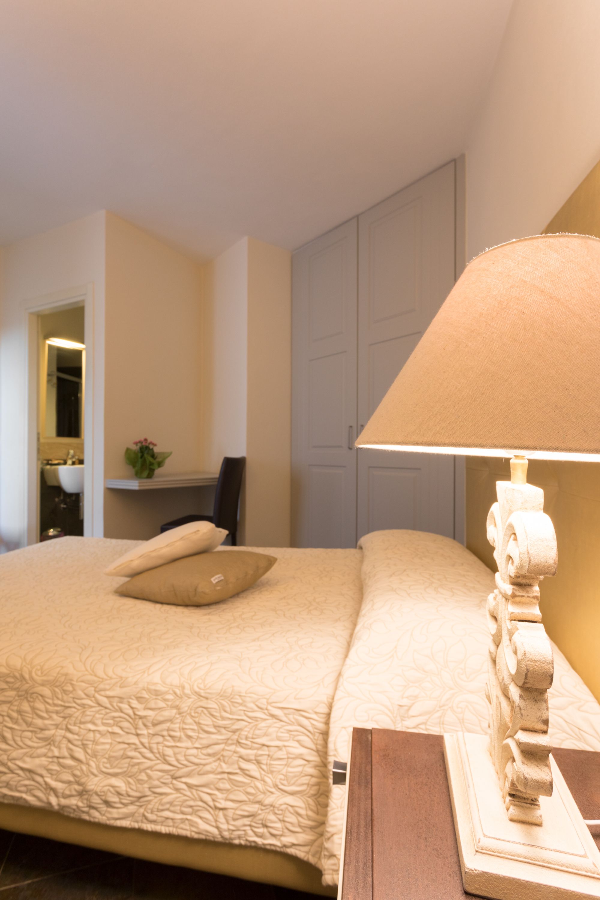 A neatly made bed with a beige quilt and pillows, a wooden nightstand with a lamp, and a bathroom visible in the background.