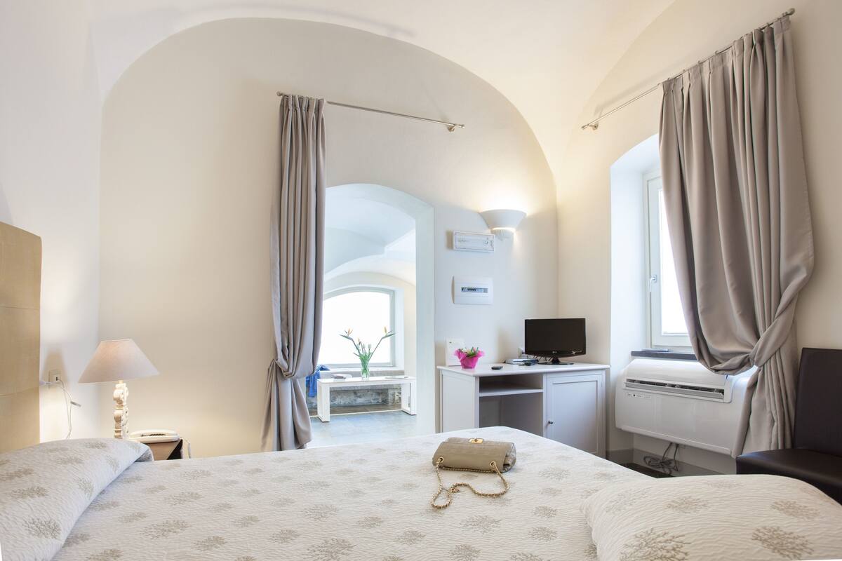 Ein Hotelzimmer mit Bett, Schreibtisch und Computer. Es gibt ein Fenster mit Vorhängen, eine Lampe und eine kleine Pflanze.
