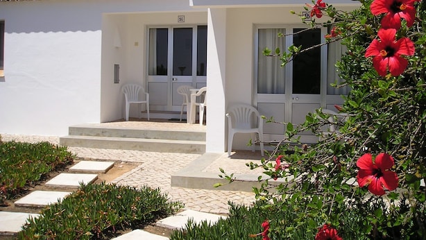 A white building with a tiled roof, a balcony with white chairs, and a red flowering plant in the foreground.