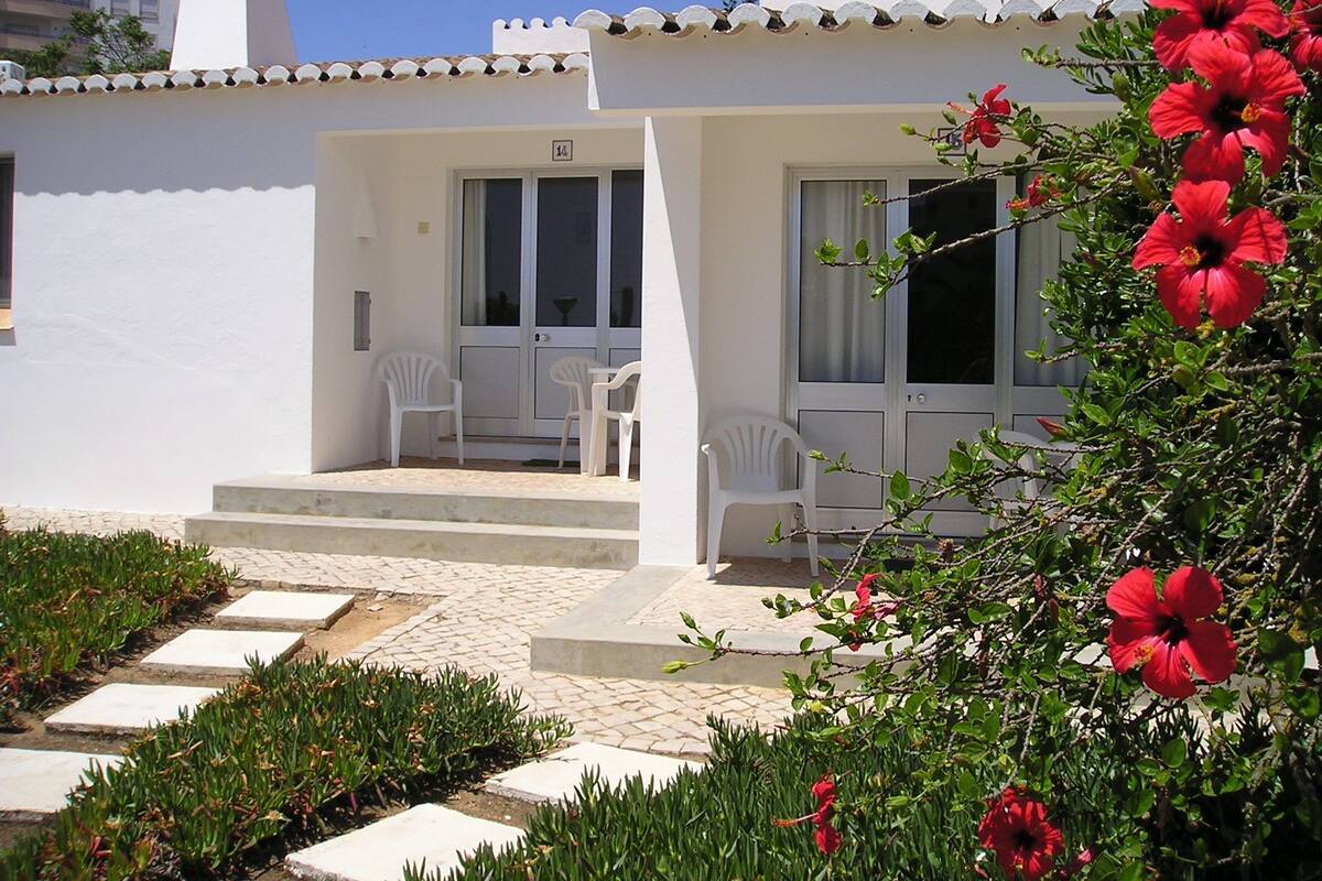 Un bâtiment blanc avec un toit recouvert de tuiles, un balcon avec des chaises blanches et, au premier plan, une plante à fleurs rouges.