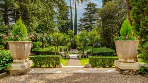 Ein formell angelegter Garten mit einem zentralen Weg, der zu einer Sitzgruppe führt, umgeben von ordentlich geschnittenen Hecken und hohen Bäumen.