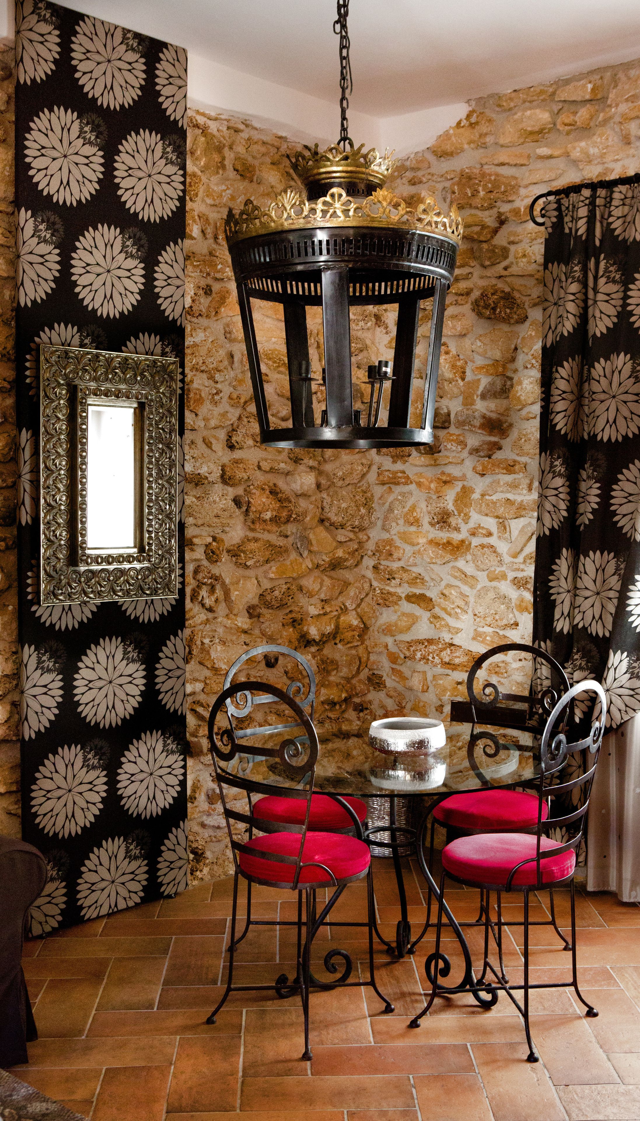 A dining area with a stone wall, a chandelier, a mirror, and red upholstered chairs.