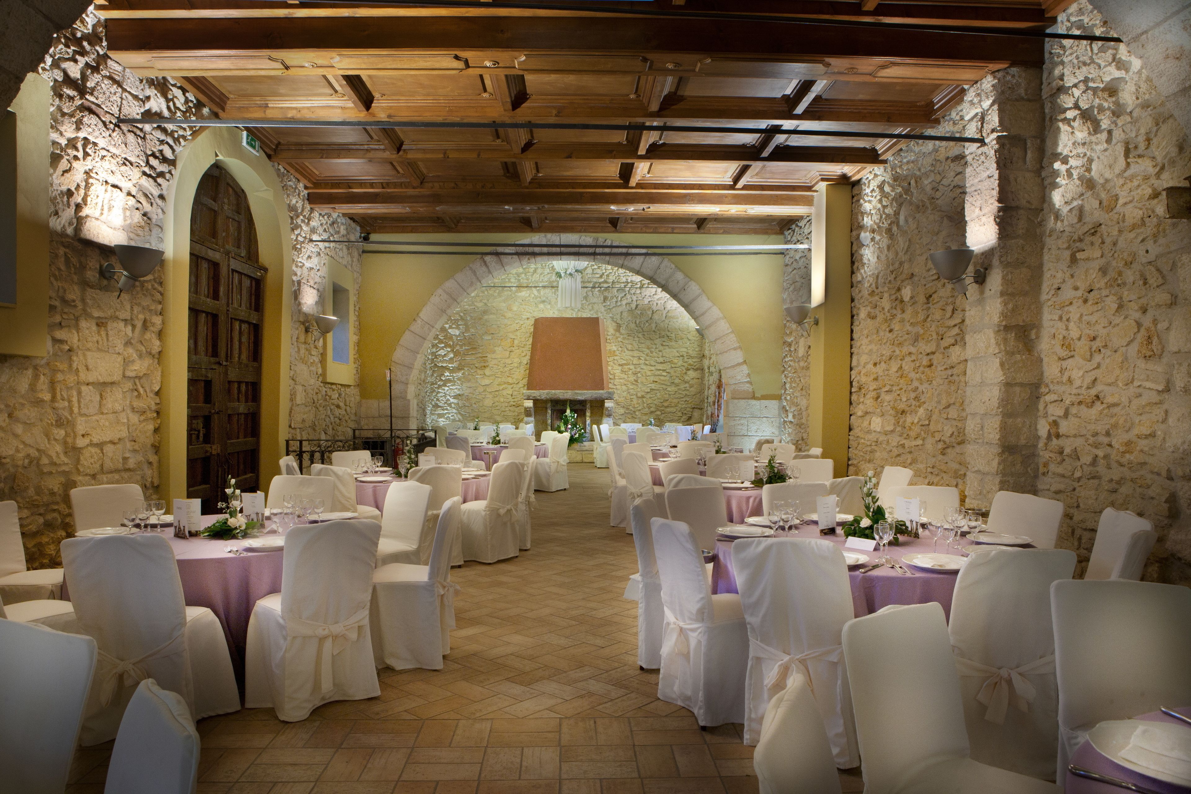 Un salón de banquetes con mesas redondas, sillas blancas y paredes de piedra.