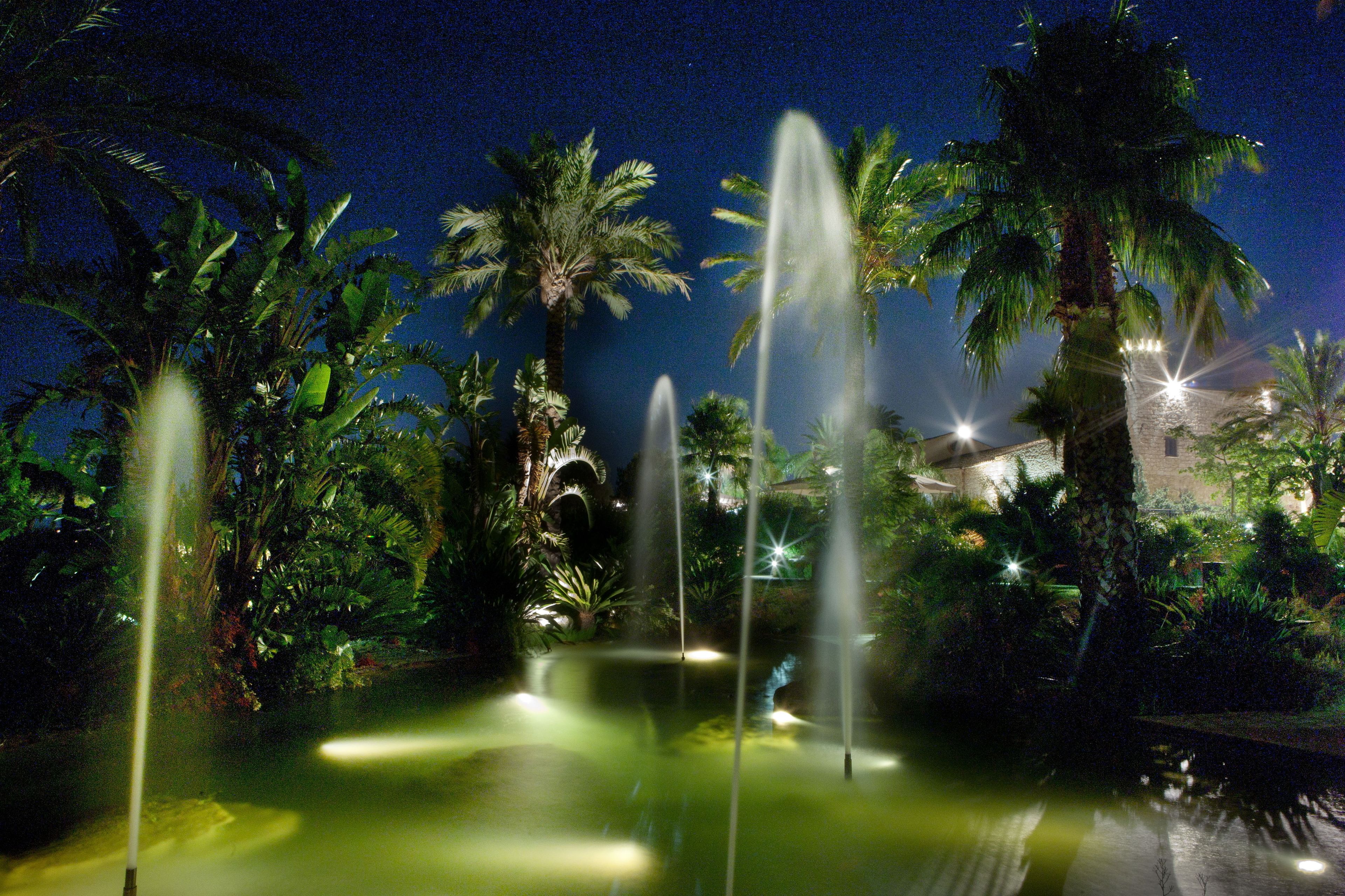 A nighttime garden with water fountains, palm trees, and a building in the background.