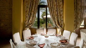 Une salle à manger avec une table dressée pour huit personnes, des chaises blanches et une vue sur l’extérieur par une fenêtre ornée de rideaux.