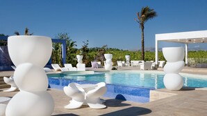 Un espace aménagé au bord de la piscine, avec des meubles blancs au design moderne, une piscine à l’eau d’un bleu limpide et une vue sur des palmiers et des montagnes.
