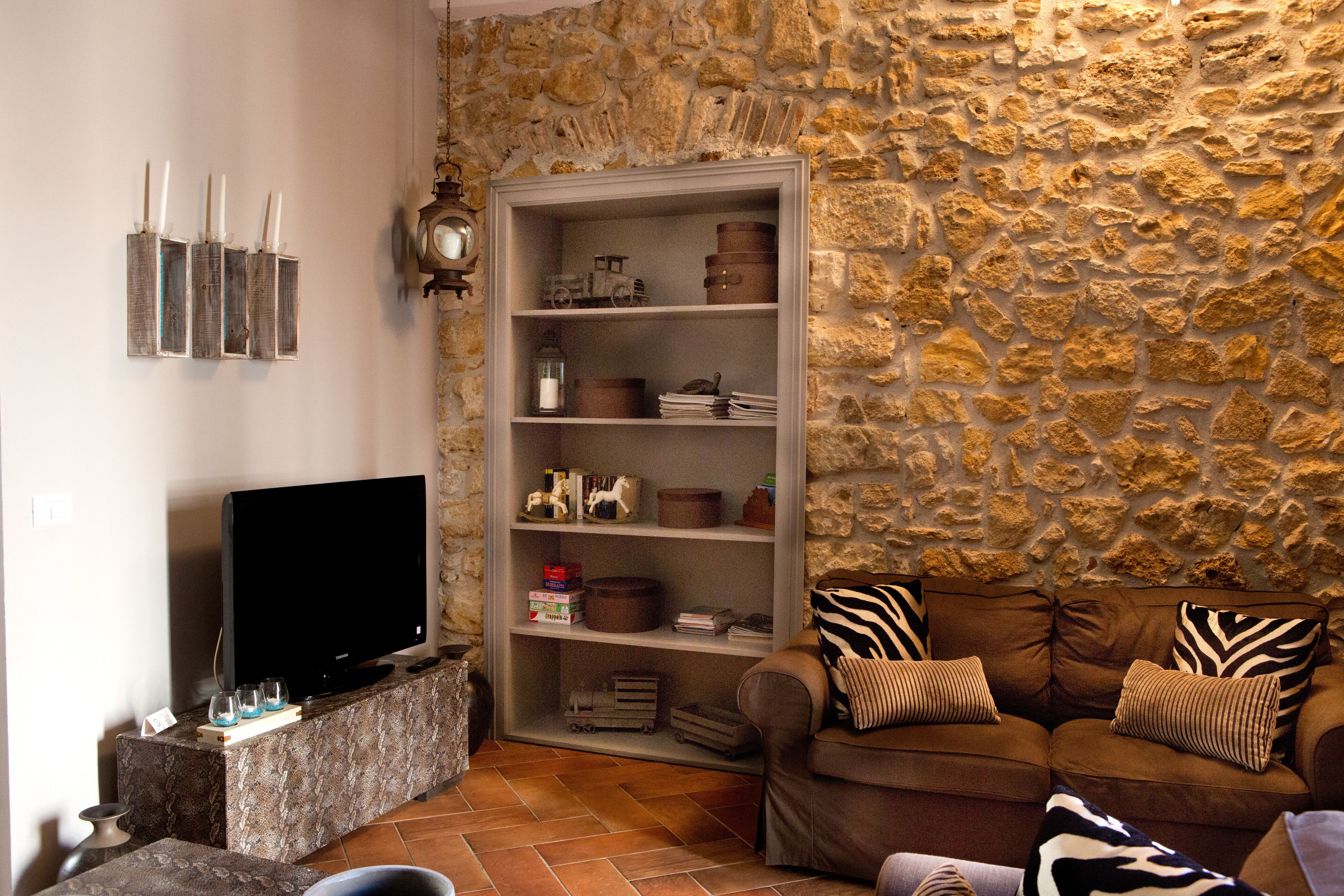 Un soggiorno con parete in pietra, una televisione su un mobile, una libreria e un divano marrone con cuscini decorati.