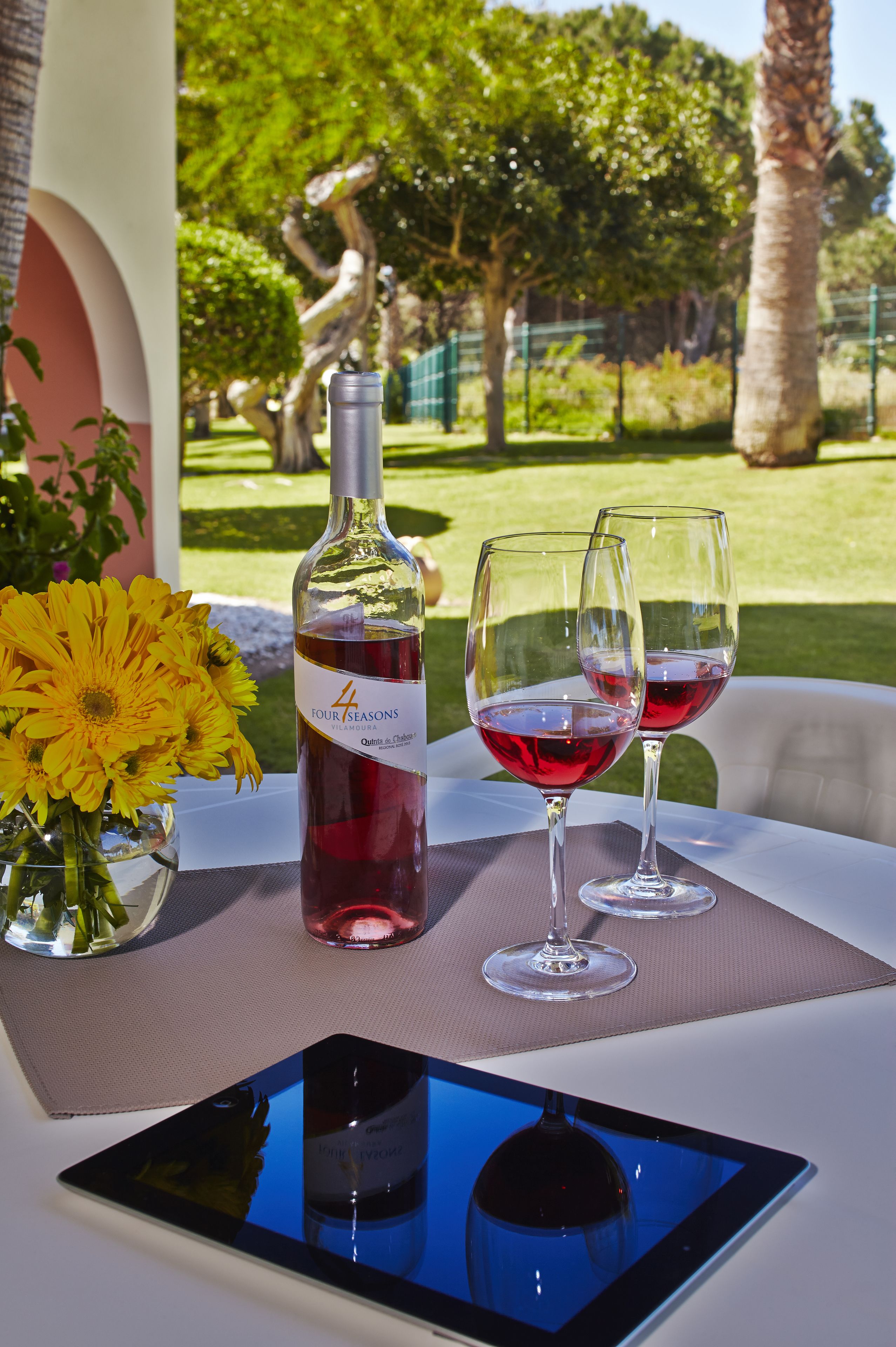 Una botella de vino, dos copas de vino, una tableta y un jarrón con girasoles sobre una mesa con vistas a un jardín.