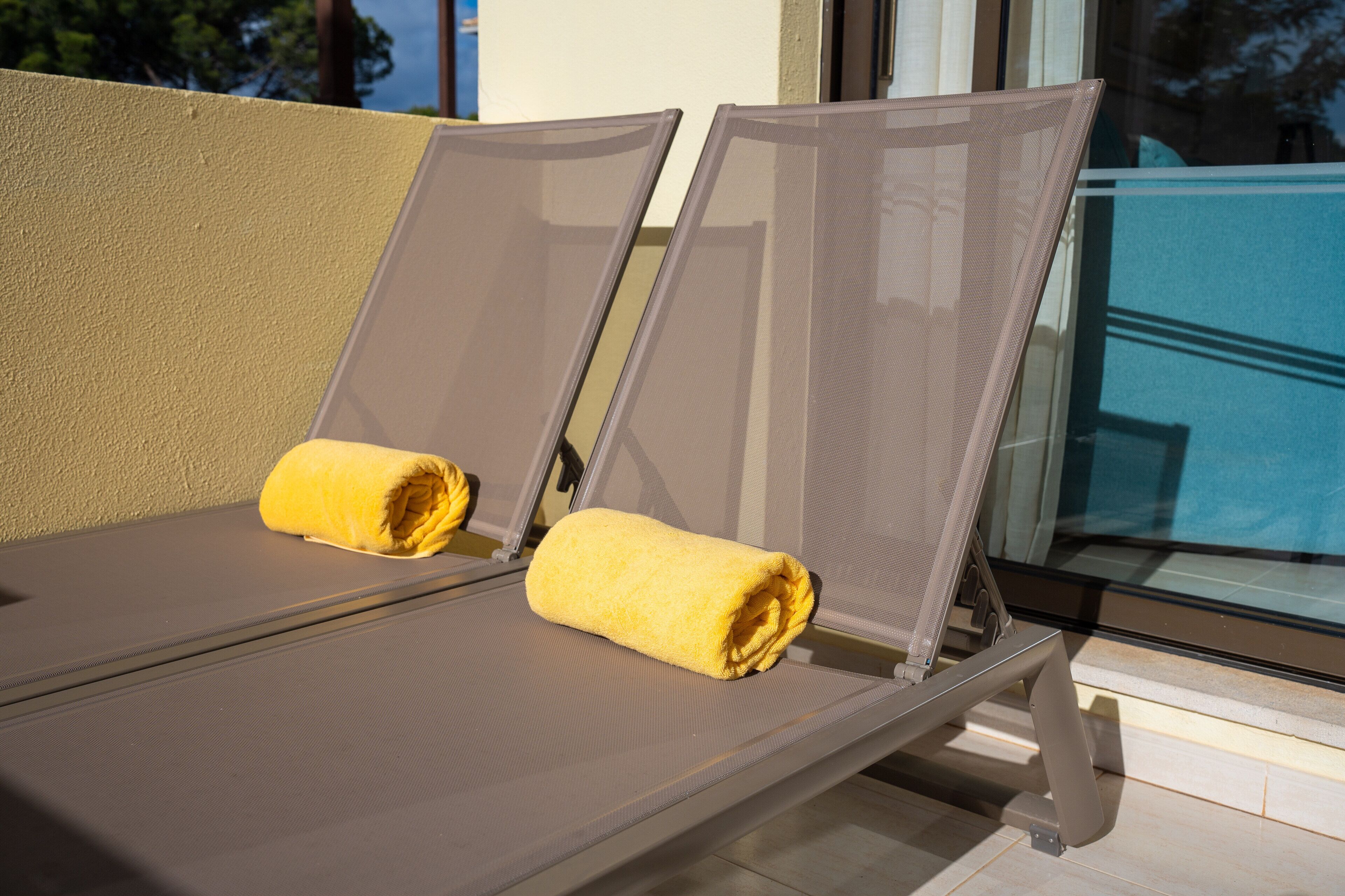 Two sun loungers with yellow towels, positioned near a glass window overlooking a pool.