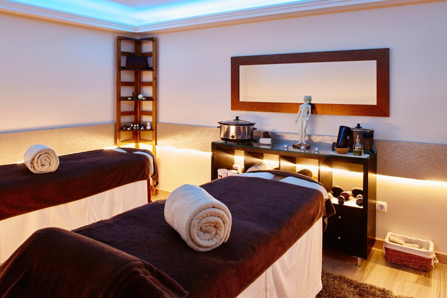A spa room with two massage tables, a shelf with toiletries, a mirror, and a small table with a kettle and a statue.