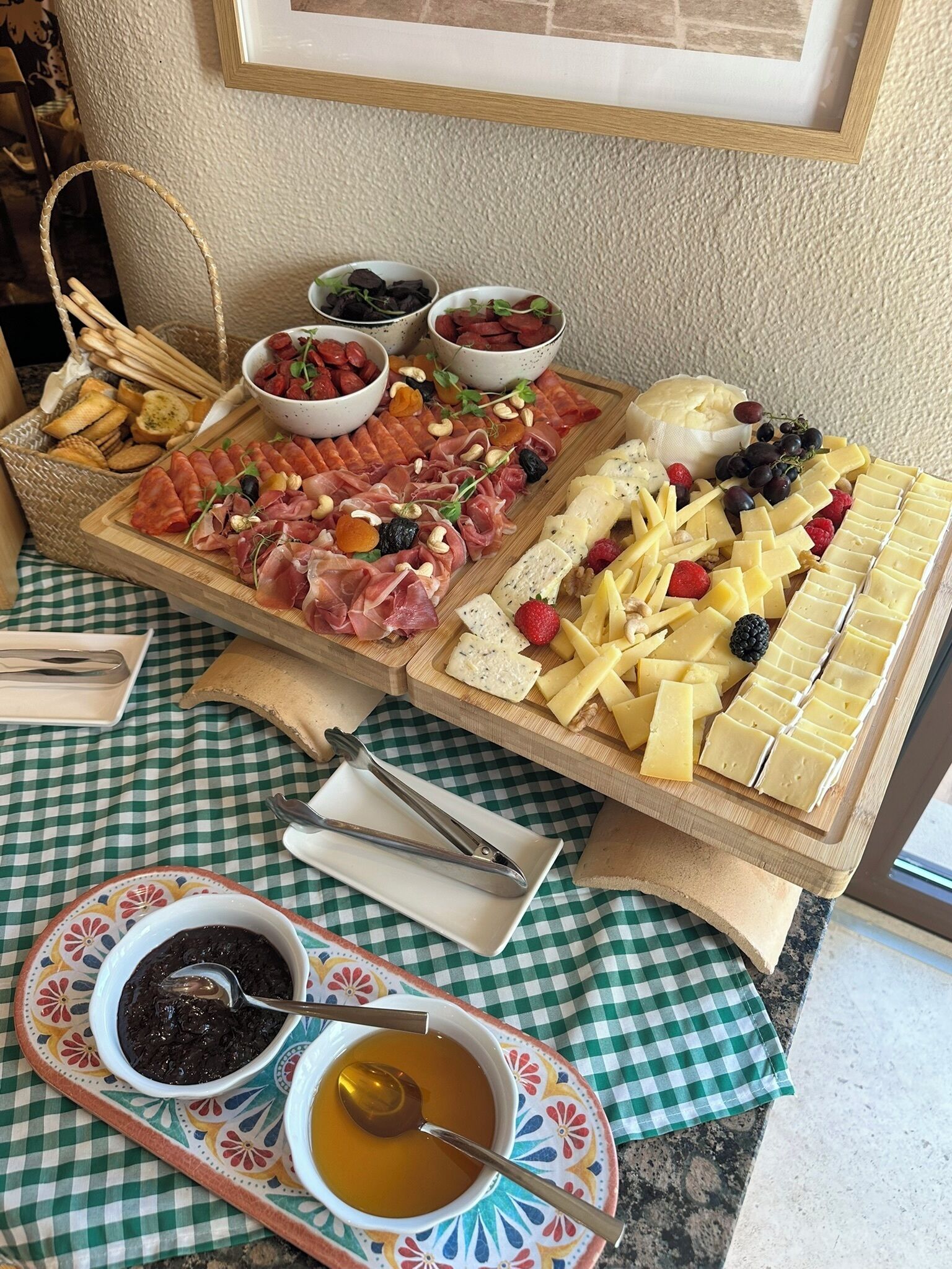 Una tabla de embutidos con varios quesos, carnes y frutas sobre un mantel a cuadros.