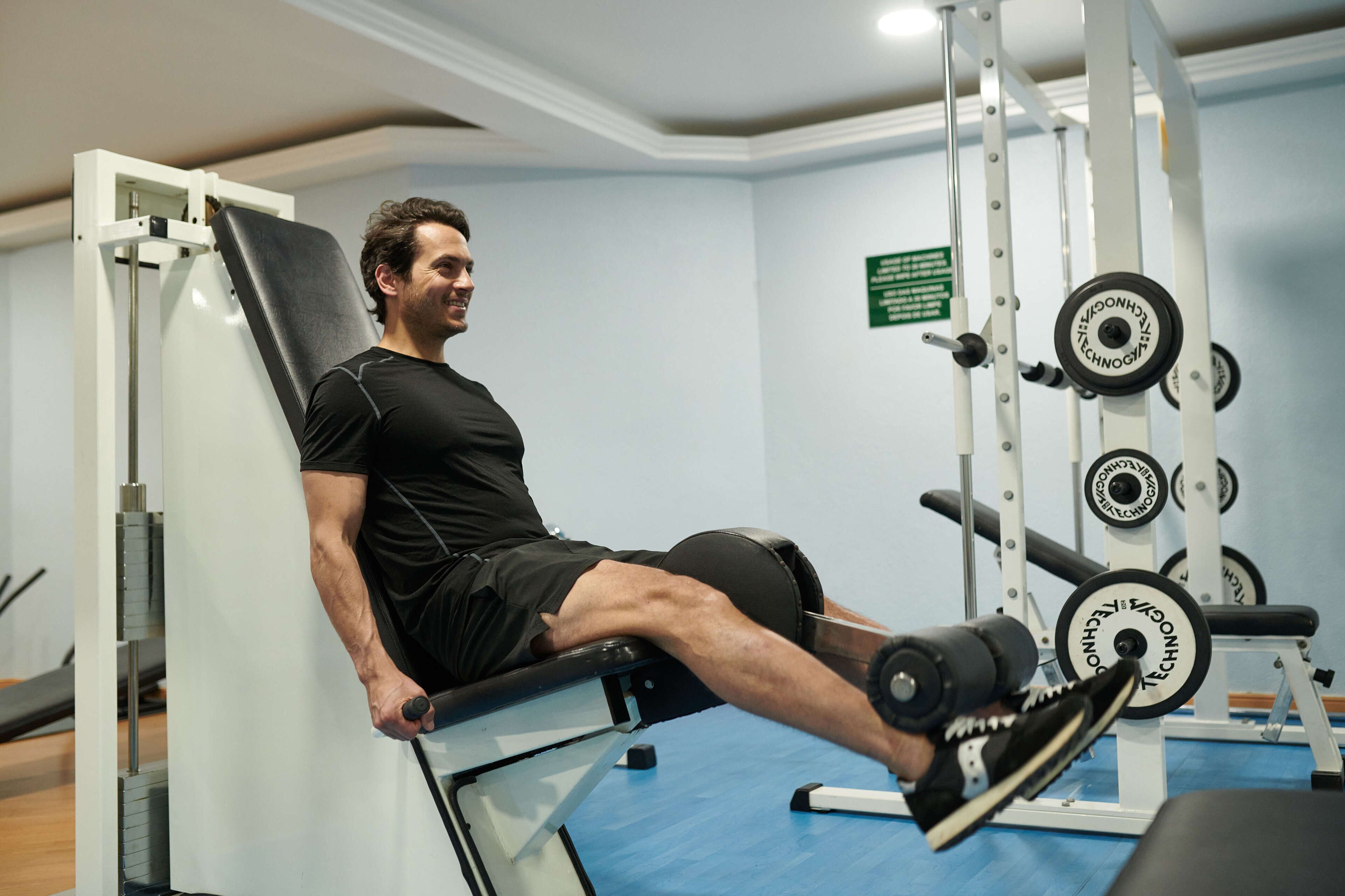Un homme utilise un appareil de musculation pour les jambes dans une salle de sport, avec des poids et un panneau vert sur le mur.