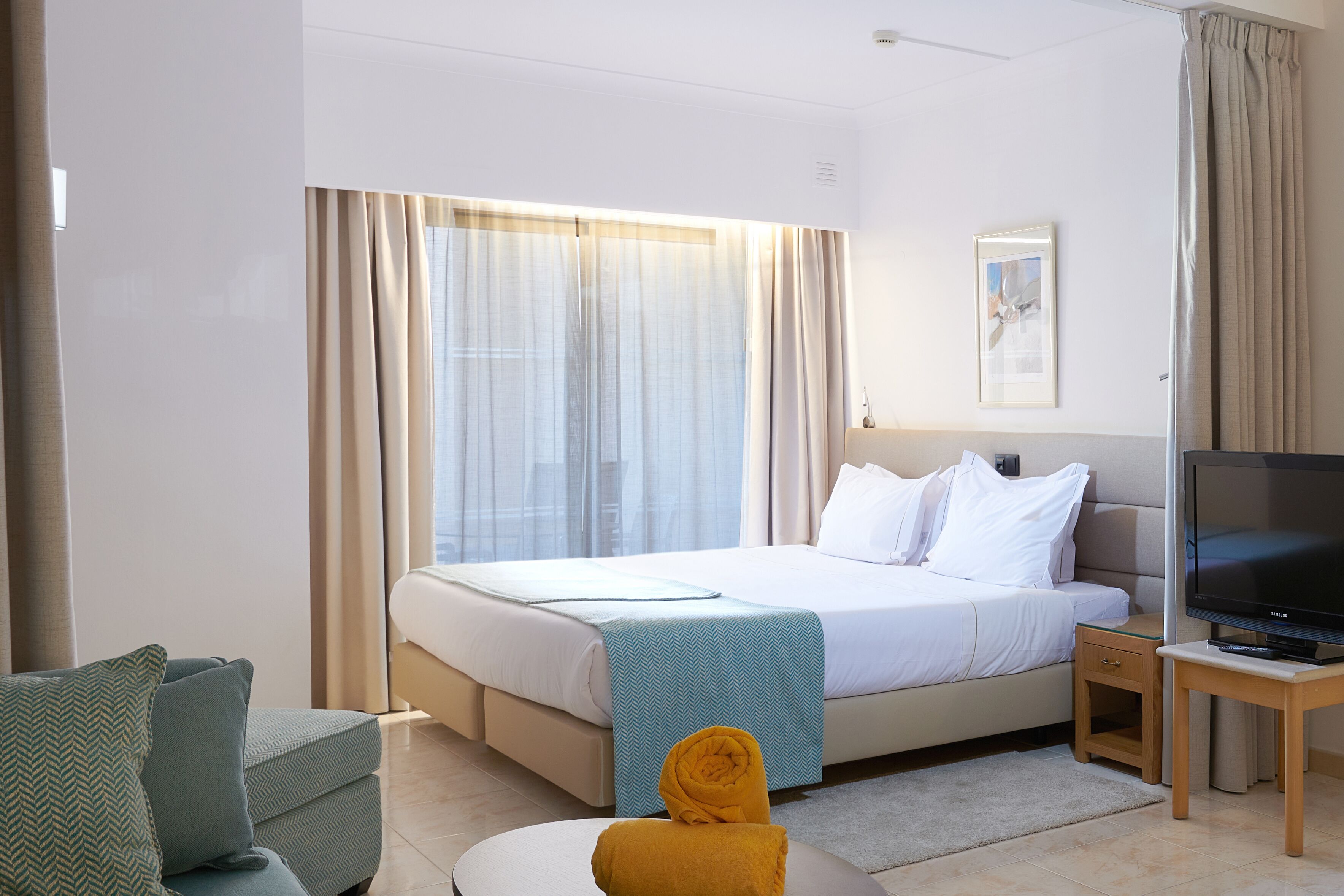 Een hotelkamer met een bed, een televisie, een tafeltje, een bank en een raam met gordijnen.
