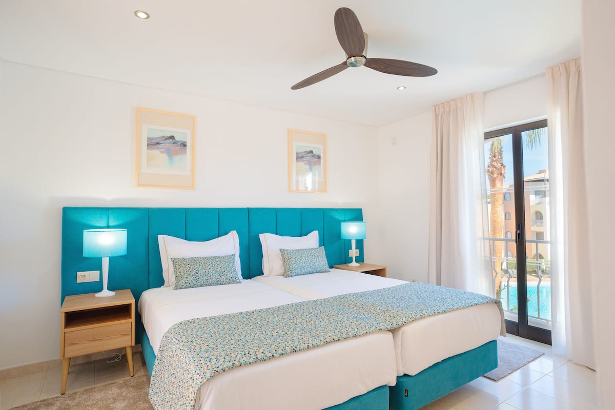 A hotel room with a large bed, two bedside tables with lamps, a ceiling fan, and two framed pictures on the wall.