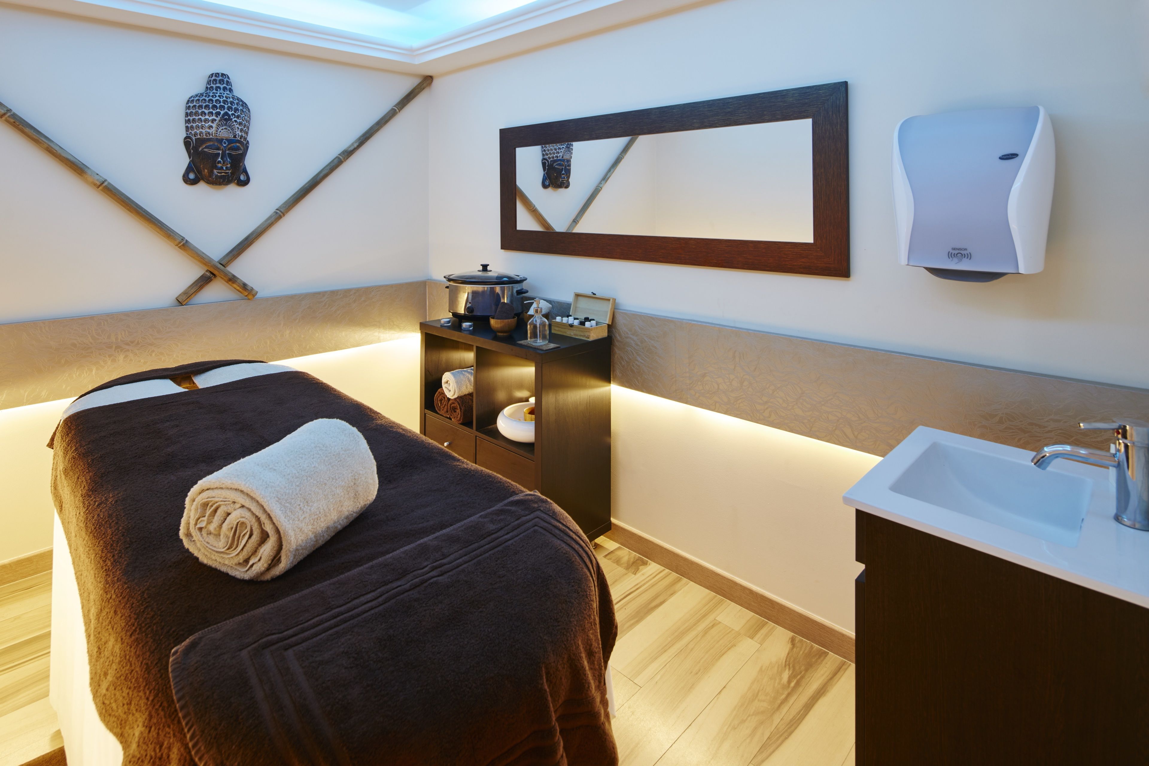 Ett spa-rum med ett massagebord, ett handfat, en spegel, en Buddha-staty och en väggmonterad tvåldispenser.