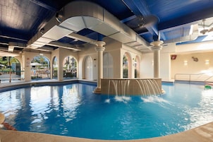 Una piscina interior con caída de agua, rodeada de columnas en arco y un techo alto con vigas a la vista.