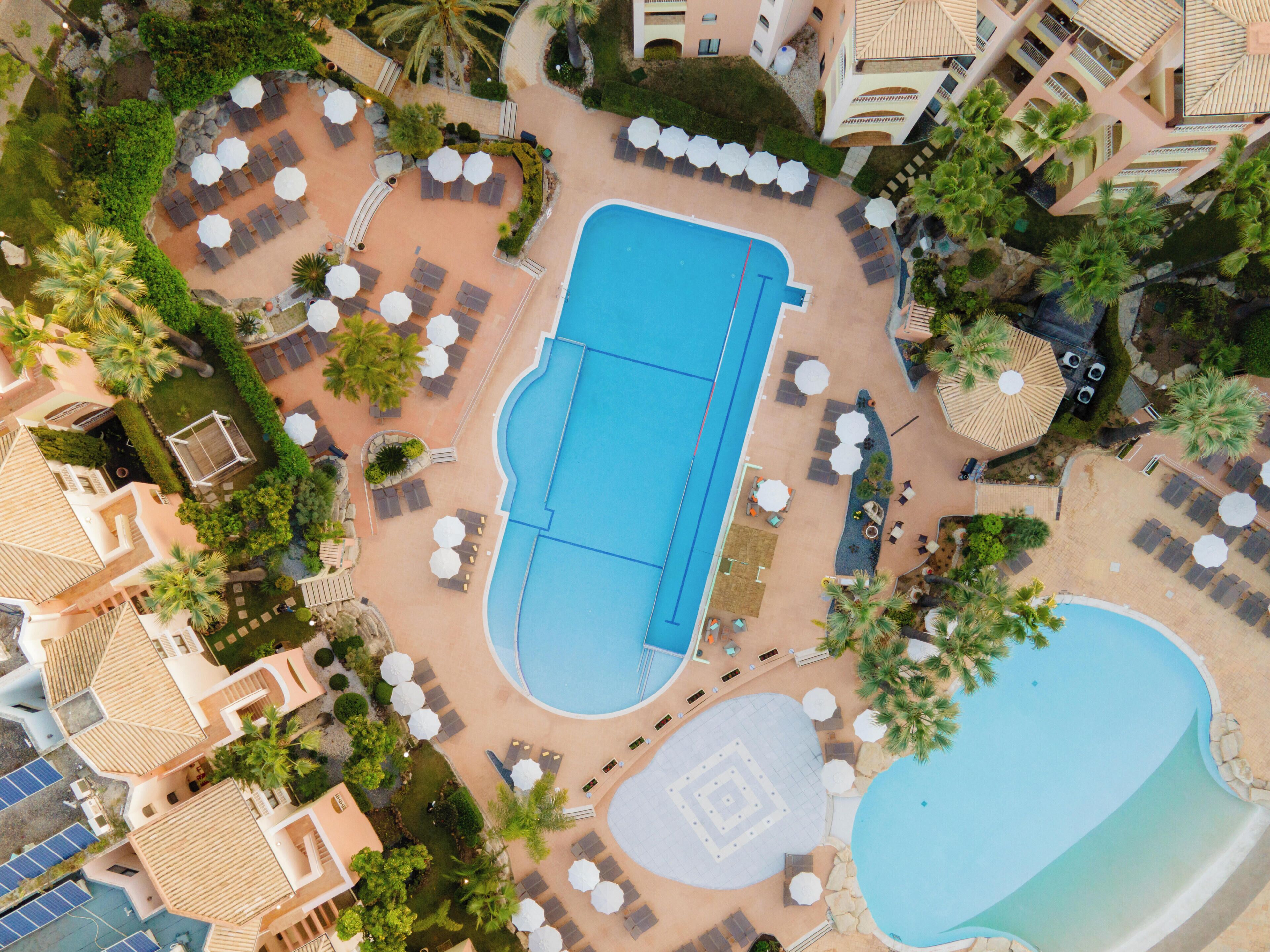 An aerial view of a resort with a large central swimming pool, multiple lounge areas with umbrellas, and surrounding palm trees.