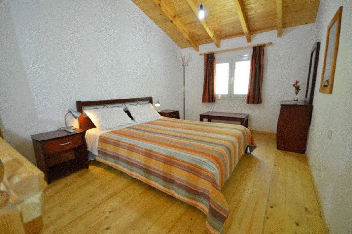 A bedroom with a wooden ceiling, a bed with a striped bedspread, two bedside tables, a nightstand, a floor lamp, a dresser, and a window with curtains.