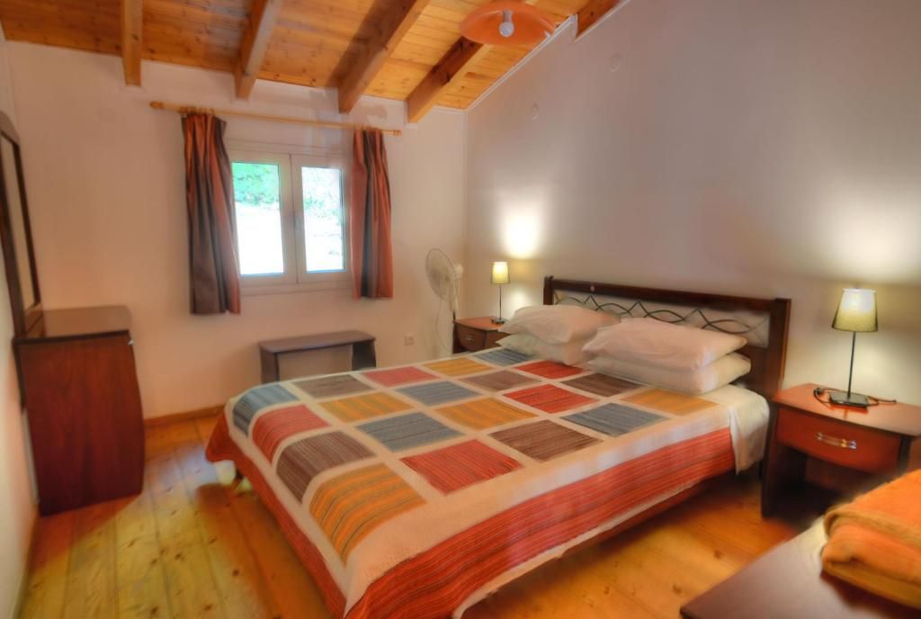 Une chambre avec un plafond en bois, un lit recouvert d’une couverture aux couleurs vives, une table de chevet avec une lampe et une fenêtre avec des rideaux.