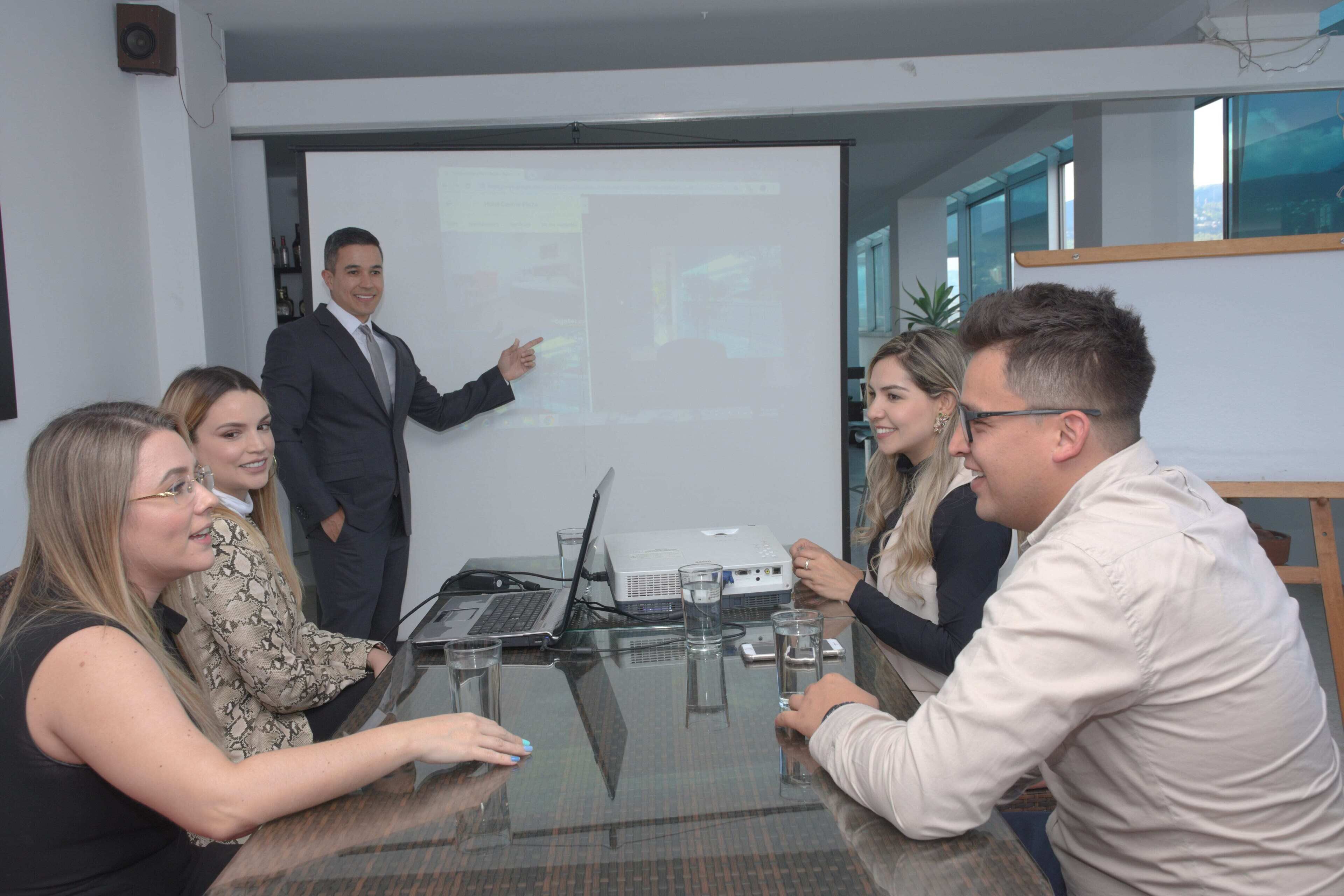 A business meeting in a modern office with a glass table, laptops, and a presentation screen.