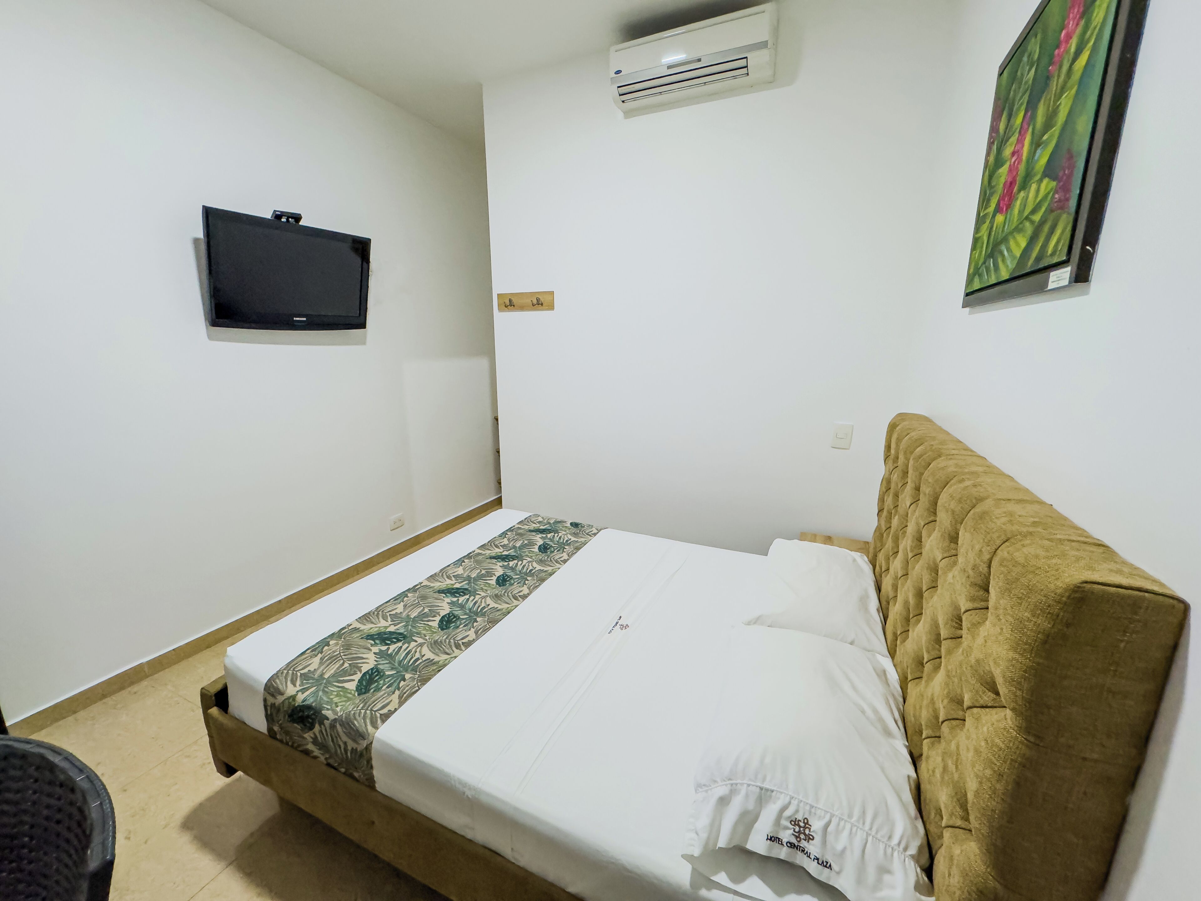 Un lit simple avec une couvre-lit à motifs floraux, un téléviseur fixé au mur et un climatiseur mural.