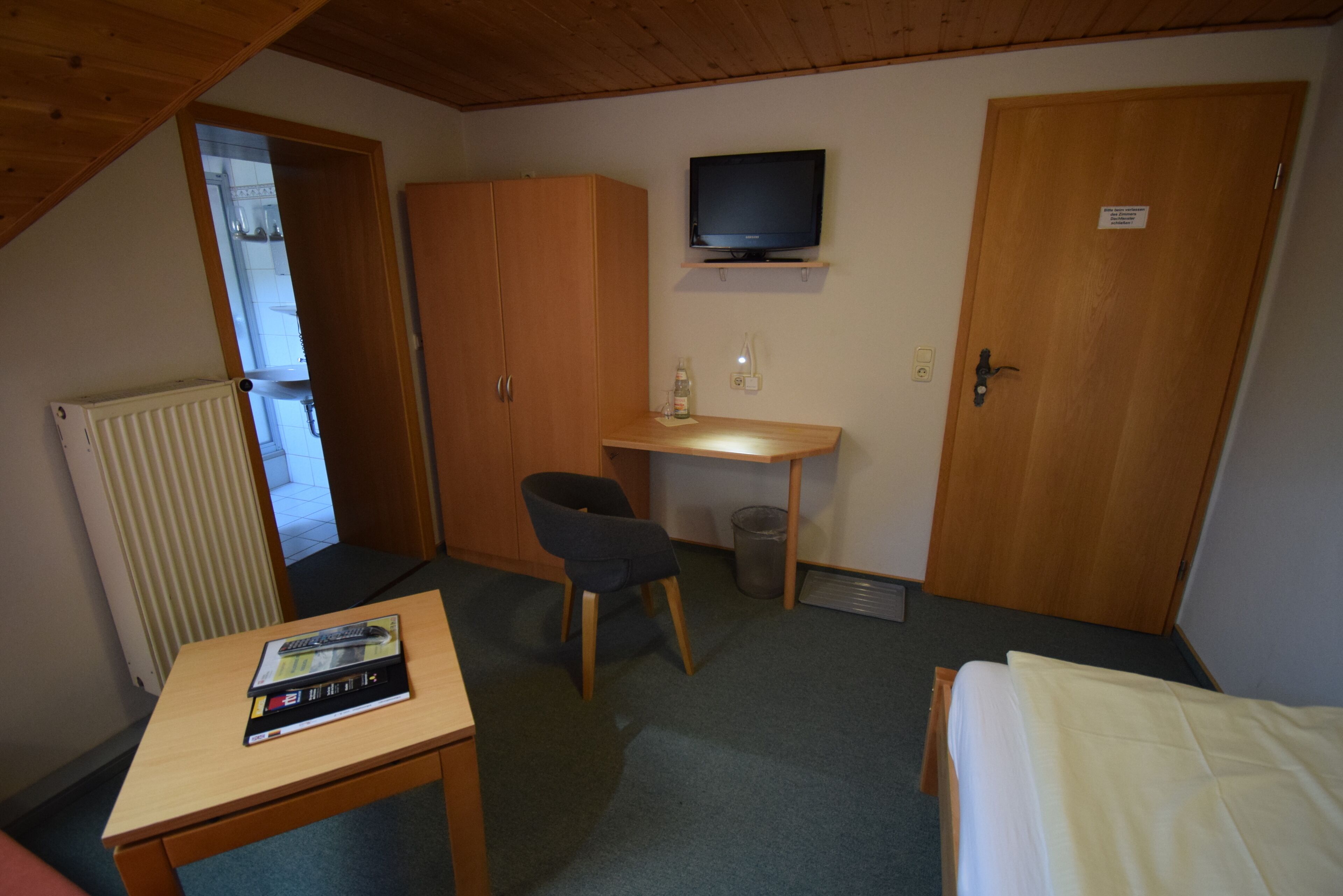 A small, furnished room with a bed, a desk, a chair, a television, and a radiator.