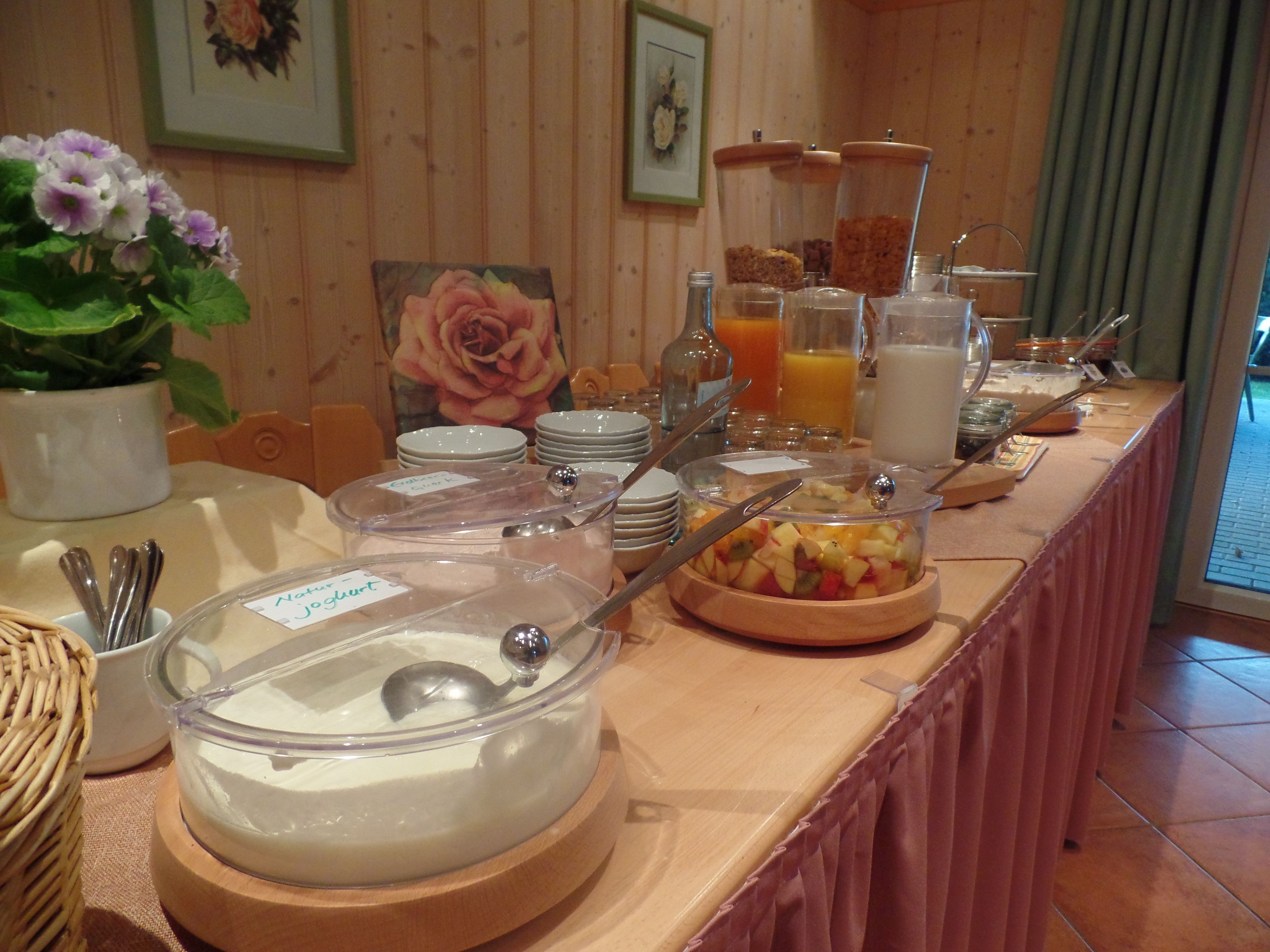 Ein reichhaltiges Buffet mit verschiedenen Speisen und Behältern, darunter eine Joghurt-Schüssel, einen Obstsalat und mehrere Getränkespender.