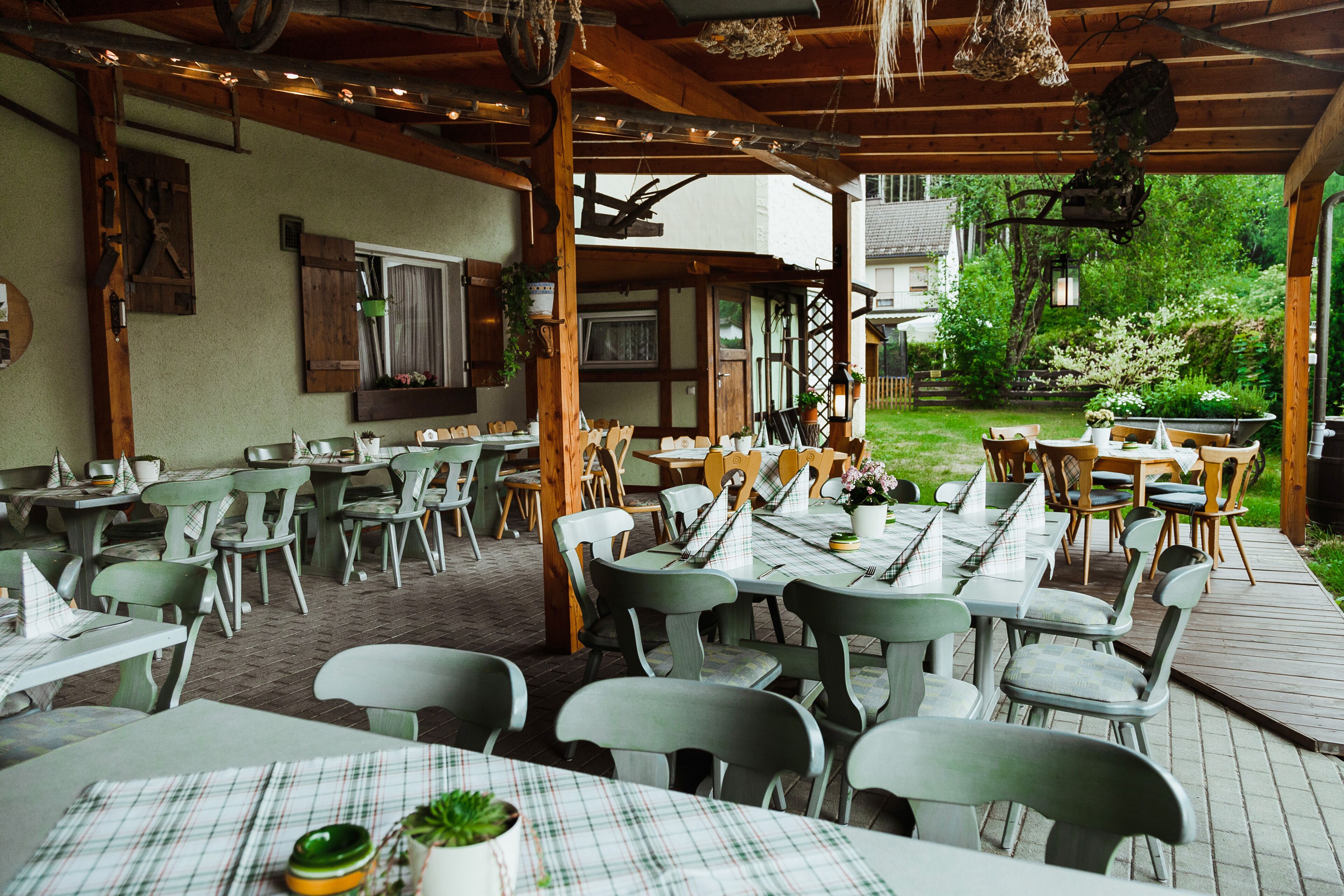 An outdoor dining area with multiple tables and chairs, wooden beams overhead, and a view of a garden and house in the background.