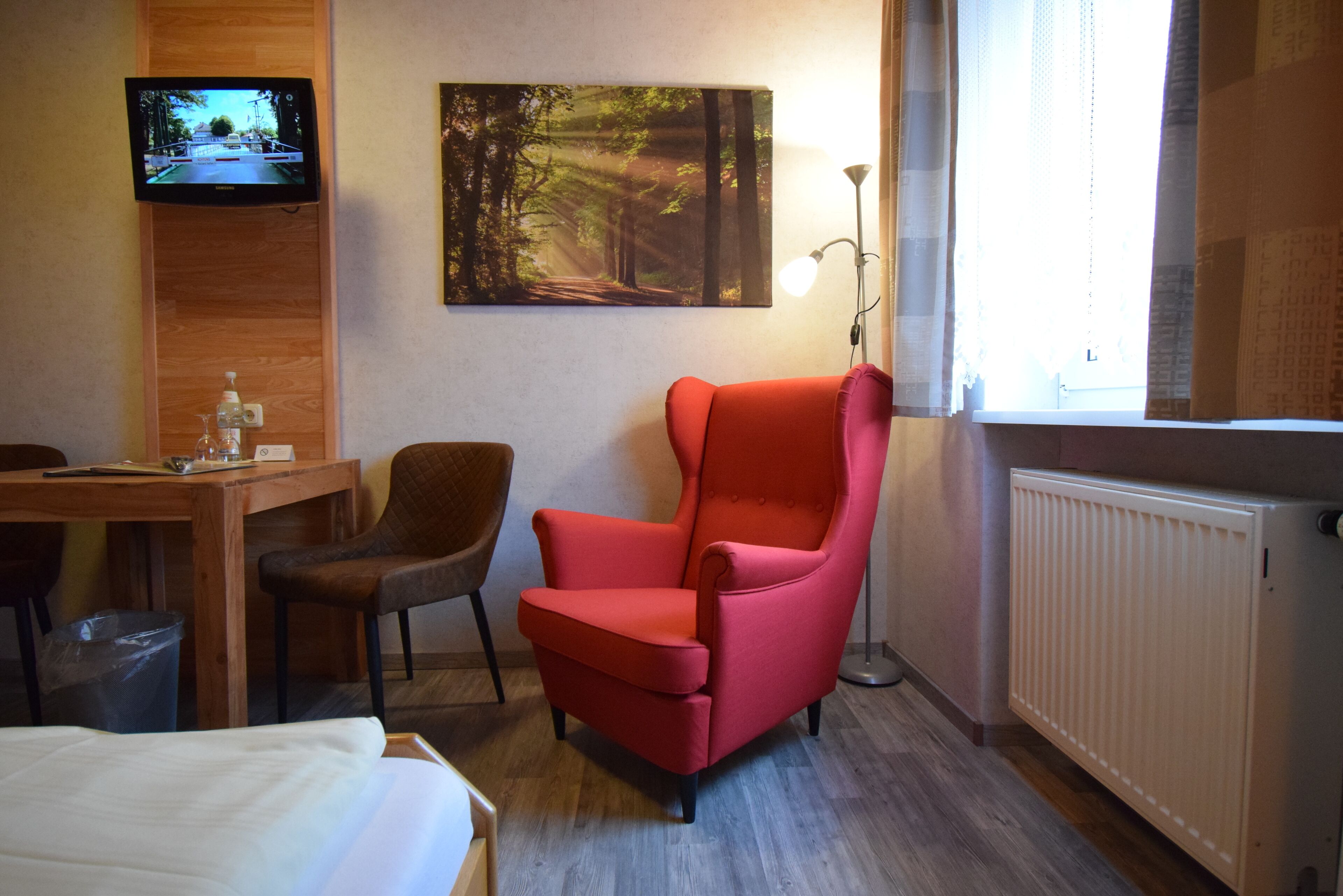 Una camera d'albergo con un letto, una scrivania con una sedia, una poltrona rossa, una TV fissata al muro, un quadro che raffigura una foresta, una lampada da terra e una finestra con tende.