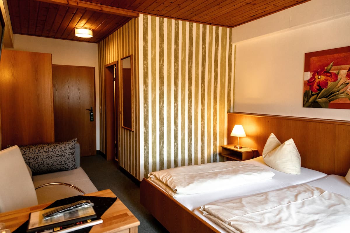 Hotelkamer met houten plafond, een gestreept scheidingswand, een bed met witte lakens, een nachtkastje met een lamp en een schilderij aan de muur.