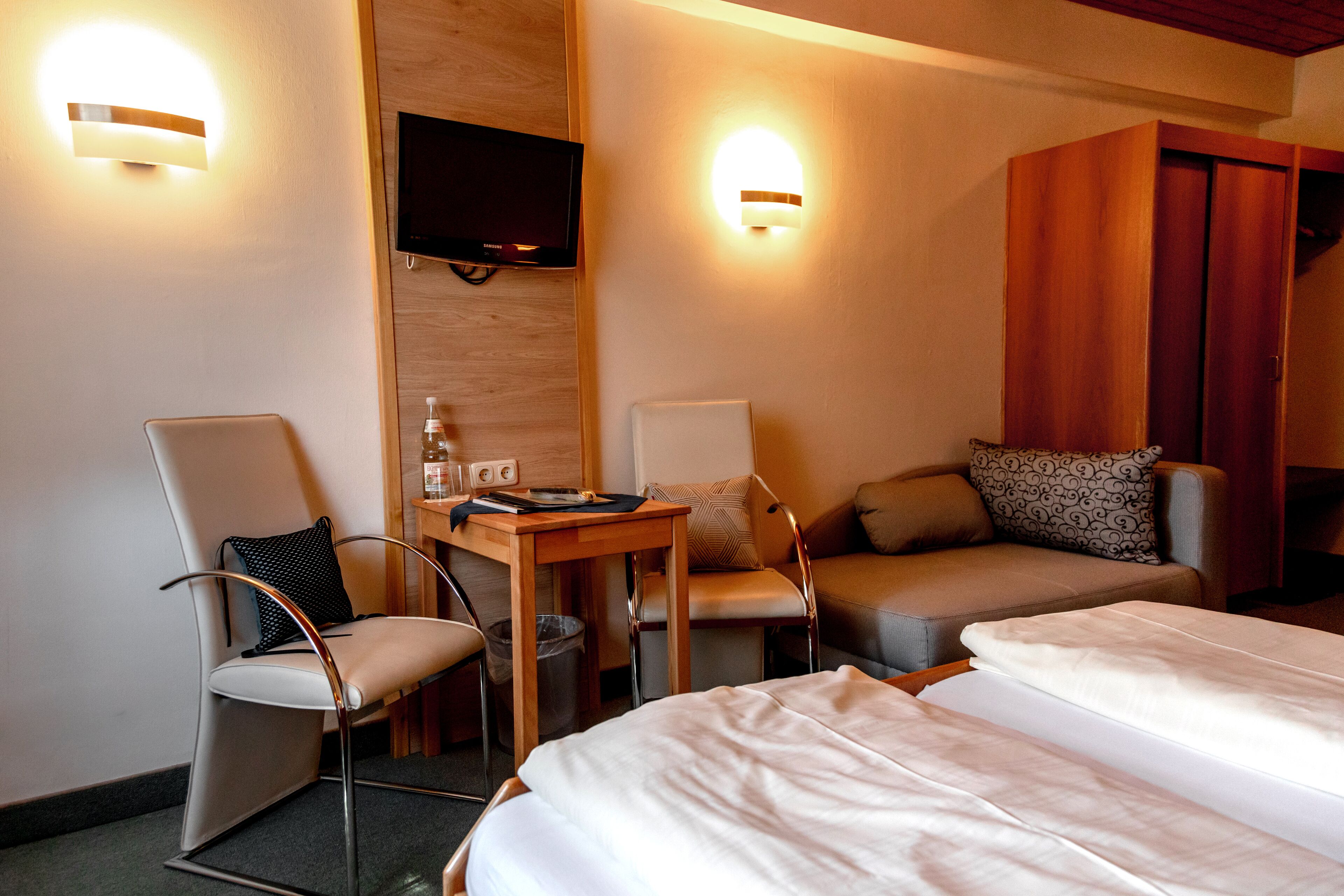 A hotel room with two single beds, a wooden desk with a chair, a television mounted on the wall, and two wall-mounted light fixtures.