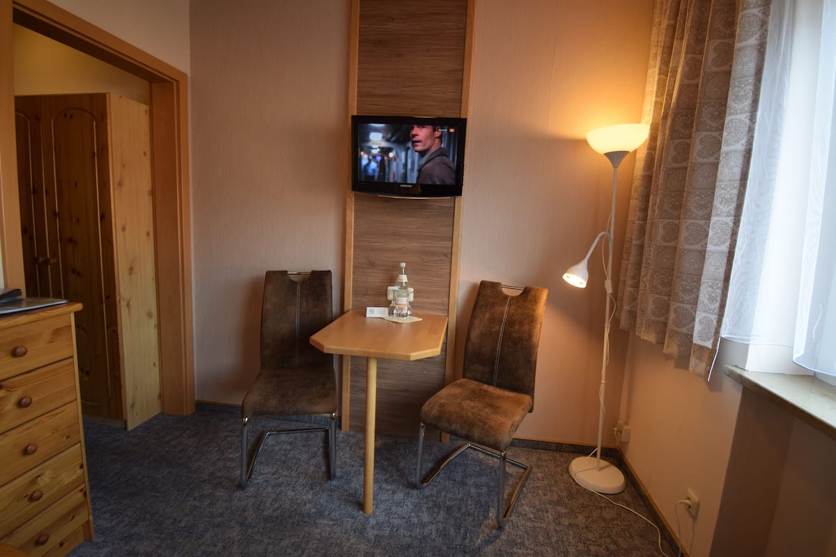 Een hotelkamer met een aan de muur bevestigde televisie, een tafeltje met een fles, twee stoelen, een dressoir en een vloerlamp.
