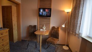 Habitación de hotel con televisión montada en la pared, una pequeña mesa con una botella, dos sillas, un tocador y una lámpara de pie.
