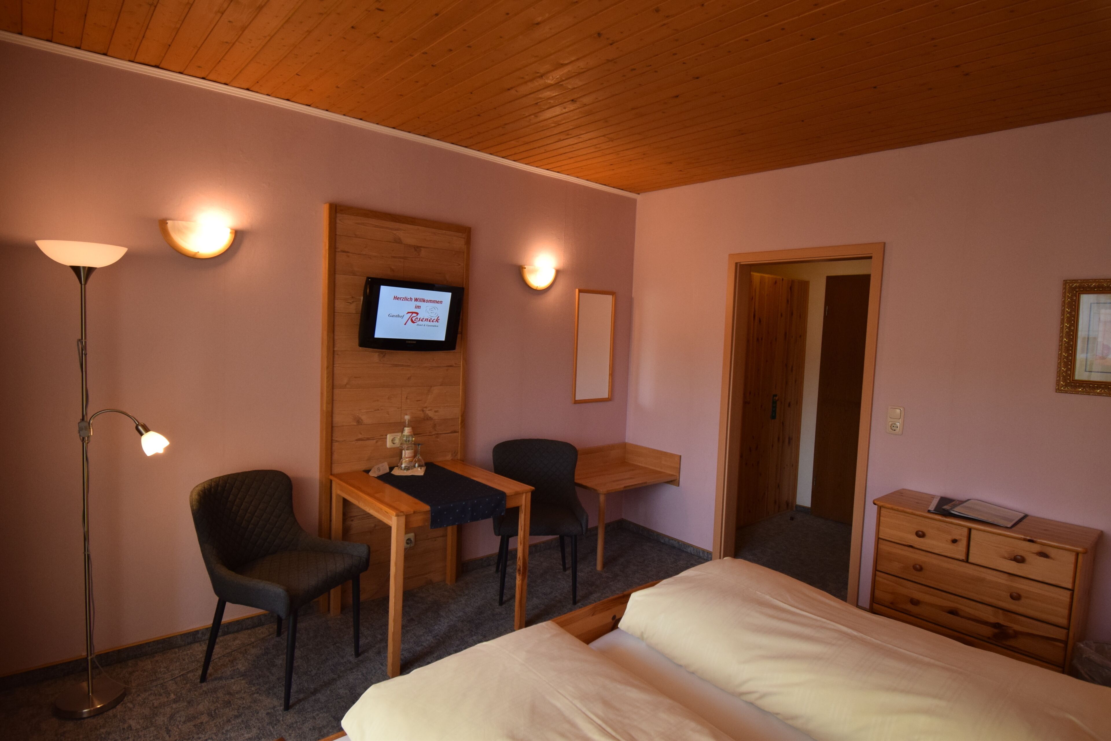 Ein Zimmer mit einem Bett, einem Schreibtisch, einem Sessel, einem Schminktisch und einem an der Wand befestigten Fernseher.