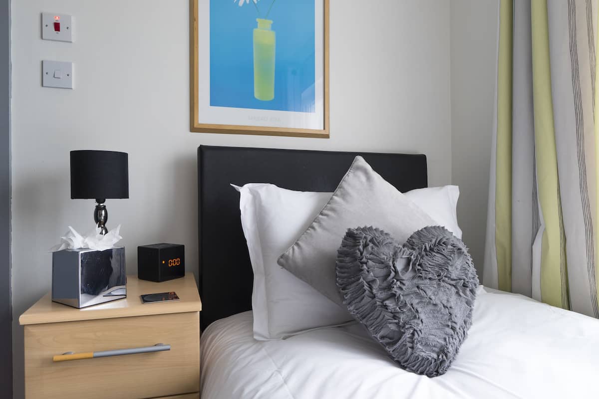 A neatly made bed with white bedding and decorative pillows, a bedside table with a lamp, a tissue box, and a digital clock, and a framed picture on the wall.