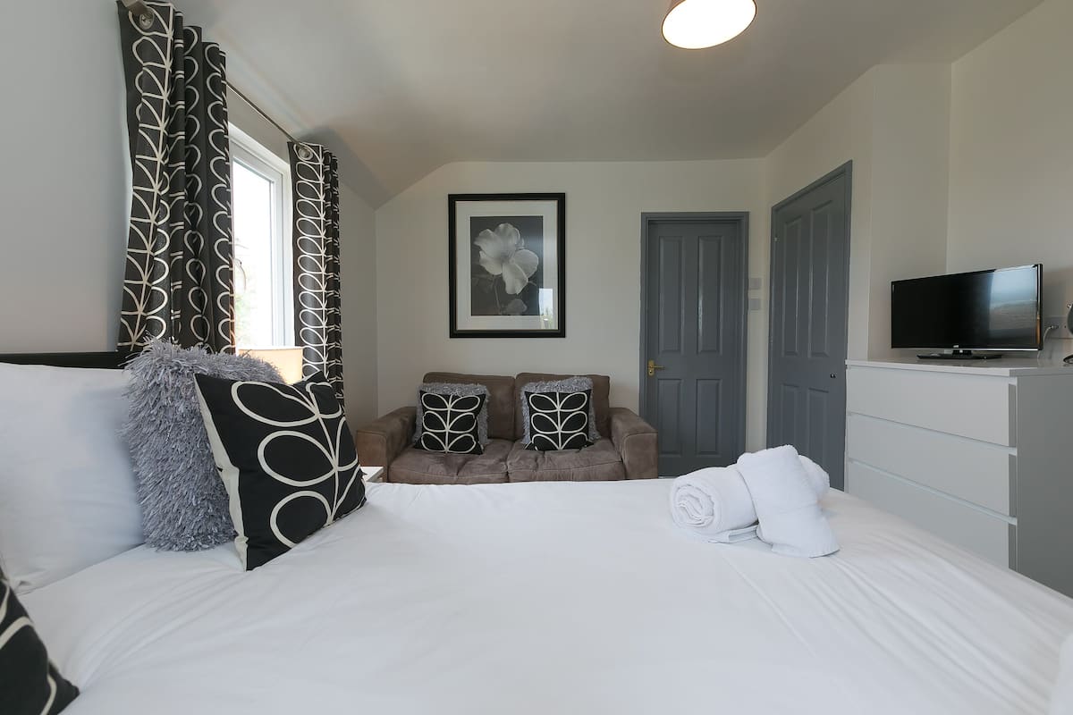 A neatly arranged bedroom with a bed, a sofa, a television, and a door.