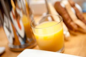A glass of orange juice on a table with baguettes and a bottle in the background. - The Nook (St Ives)