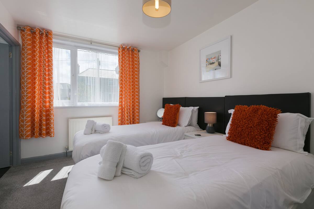 A hotel room with two beds, white bedding, orange accents, a framed picture on the wall, and a window with curtains.